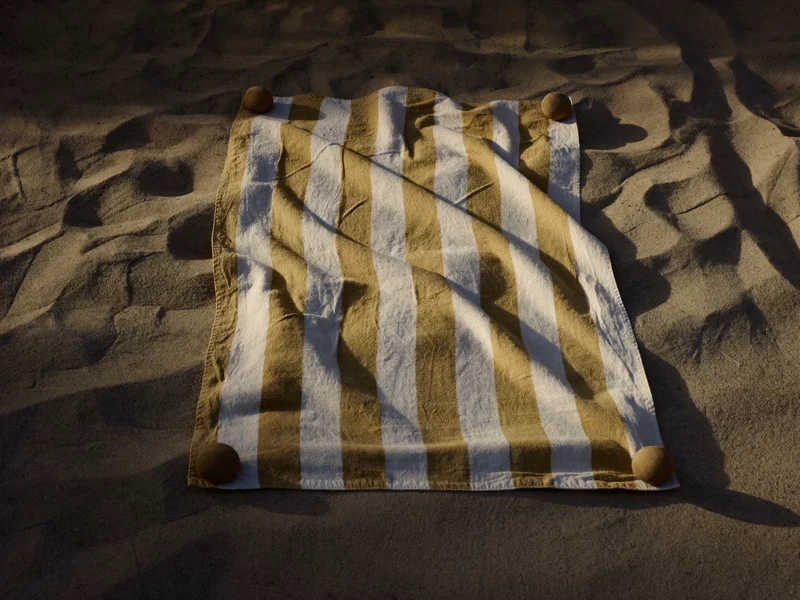Gestreiftes Leinen-Baumwoll-Strandtuch auf hellem Sand mit Kieselsteinen an den Ecken